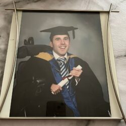 Martyn dressed in his graduation outfit in 2006 with a black hat, gown, blue and white tie, and a gold and blue sash is sat smiling to the camera holding a paper scroll
