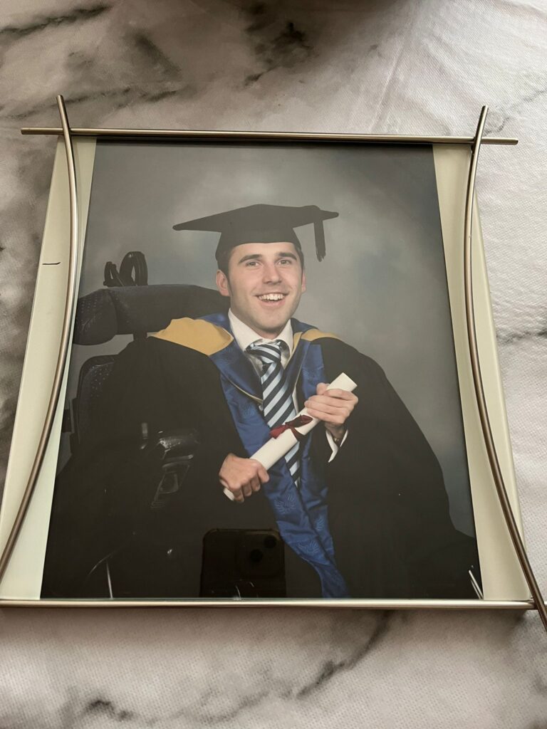 Martyn dressed in his graduation outfit in 2006 with a black hat, gown, blue and white tie, and a gold and blue sash is sat smiling to the camera holding a paper scroll