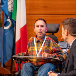 Martyn in his orange and red checked shirt with a microphone from his ear to mouth in his wheelchair talking on stage in San Marino