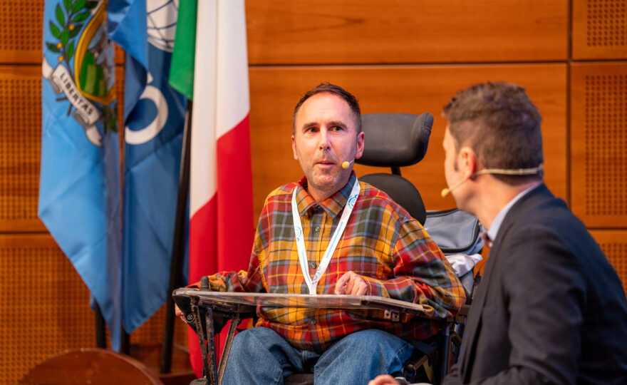 Martyn in his orange and red checked shirt with a microphone from his ear to mouth in his wheelchair talking on stage in San Marino
