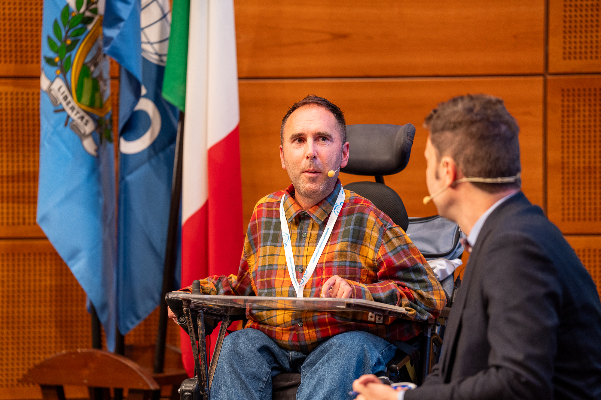 Martyn in his orange and red checked shirt with a microphone from his ear to mouth in his wheelchair talking on stage in San Marino 