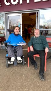 Martyn is a white male with short brown hair wearing a light blue hoody, grey trousers and white converse shoes, he's sat in his wheelchair next to Dave a white male with short brown hair, a beard, green jumper, black trousers and shoes sat on a red chair outside a cafe