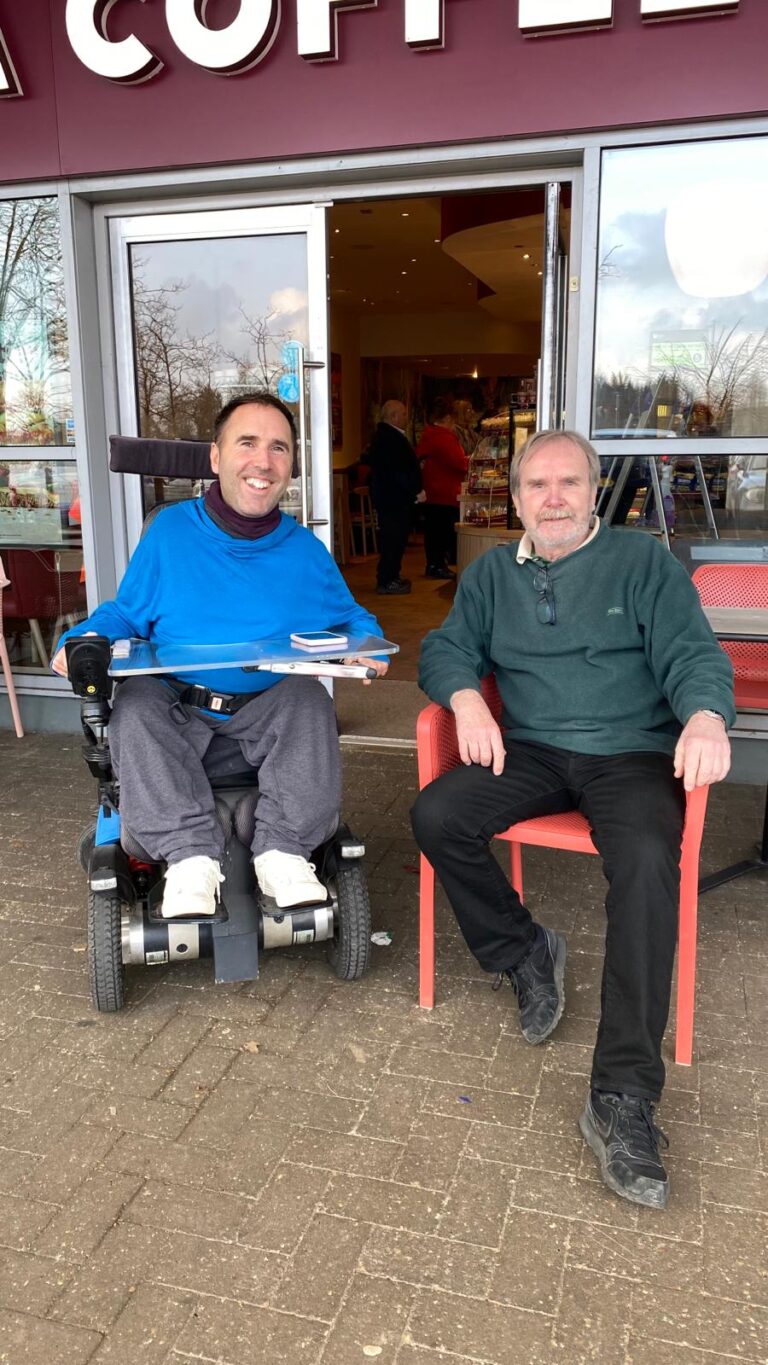 Martyn is a white male with short brown hair wearing a light blue hoody, grey trousers and white converse shoes, he's sat in his wheelchair next to Dave a white male with short brown hair, a beard, green jumper, black trousers and shoes sat on a red chair outside a cafe