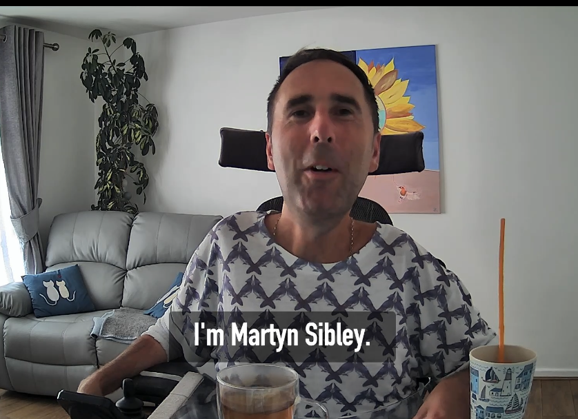 Martyn Sibley, a white male with short brown hair, is sitting in a well-lit living room. He is smiling warmly and talking to camera, seated in his wheelchair. He is wearing a patterned shirt and has a drink with a straw on the table in front of him. Behind him, there is a sofa, a plant, and a painting of a sunflower on the wall.