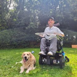 Martyn is sitting in his powered wheelchair on a grassy lawn, wearing a light grey shirt and a black cap. Beside him, his golden retriever, Sunny, lies on the grass with his tongue out, looking content. They are in an outdoor setting with tall, lush trees in the background, enjoying a sunny day together.