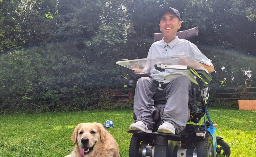 Martyn is sitting in his powered wheelchair on a grassy lawn, wearing a light grey shirt and a black cap. Beside him, his golden retriever, Sunny, lies on the grass with his tongue out, looking content. They are in an outdoor setting with tall, lush trees in the background, enjoying a sunny day together.