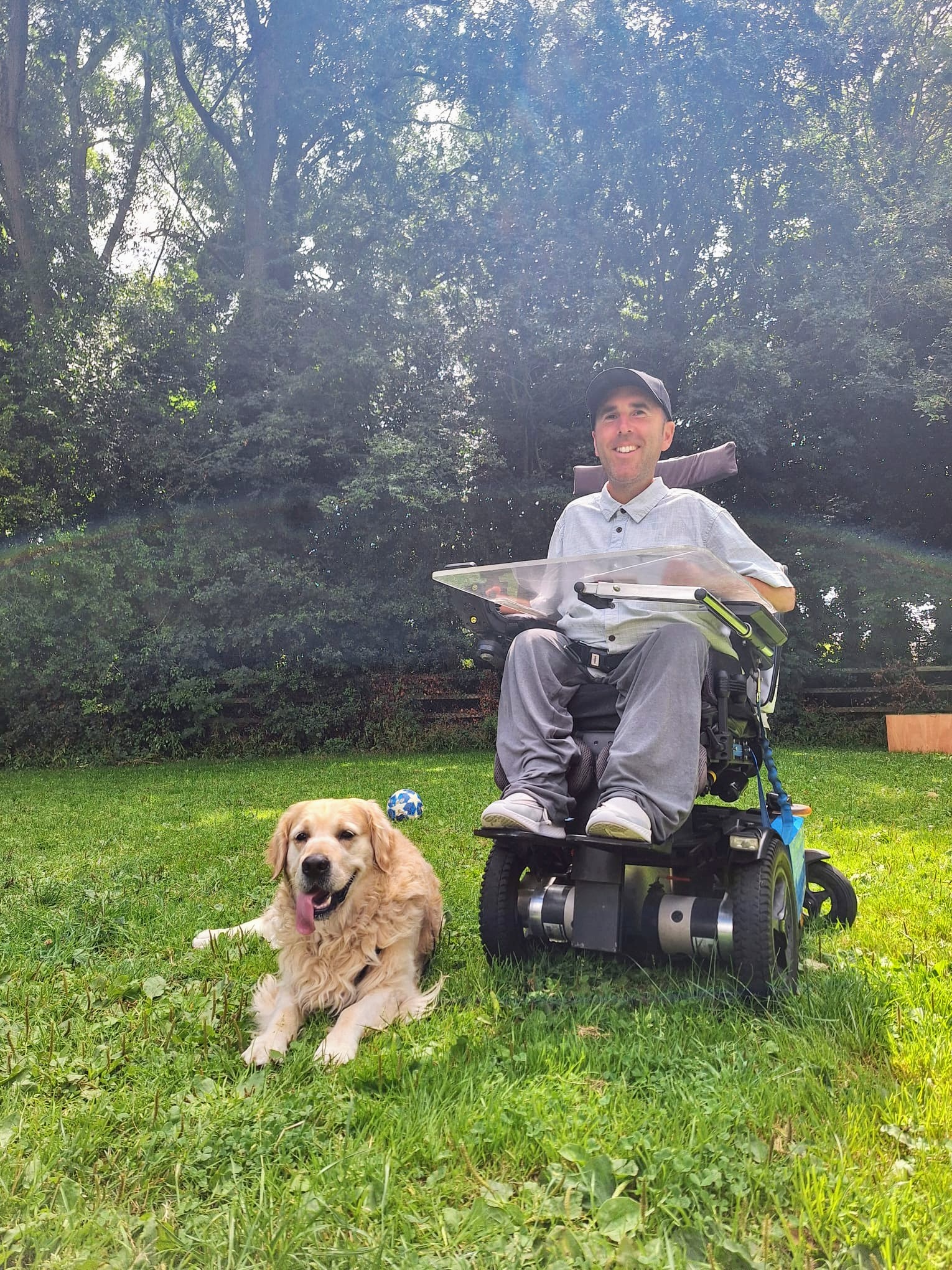 Martyn is sitting in his powered wheelchair on a grassy lawn, wearing a light grey shirt and a black cap. Beside him, his golden retriever, Sunny, lies on the grass with his tongue out, looking content. They are in an outdoor setting with tall, lush trees in the background, enjoying a sunny day together.