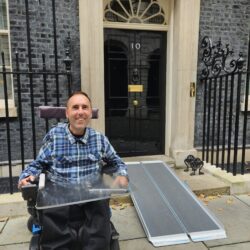 Alt text: Martyn Sibley, sitting in his wheelchair, smiles in front of the famous black door of 10 Downing Street. A ramp is laid out in front of the entrance, highlighting the building's accessibility for wheelchair users. Martyn is wearing a blue and black plaid shirt and dark trousers, with the iconic address plate and lion's head door knocker visible behind him.