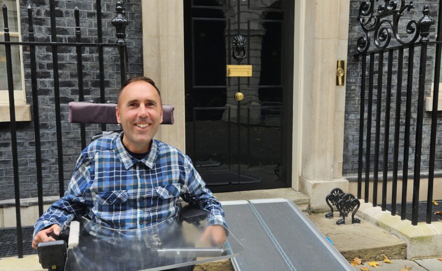 Alt text: Martyn Sibley, sitting in his wheelchair, smiles in front of the famous black door of 10 Downing Street. A ramp is laid out in front of the entrance, highlighting the building's accessibility for wheelchair users. Martyn is wearing a blue and black plaid shirt and dark trousers, with the iconic address plate and lion's head door knocker visible behind him.