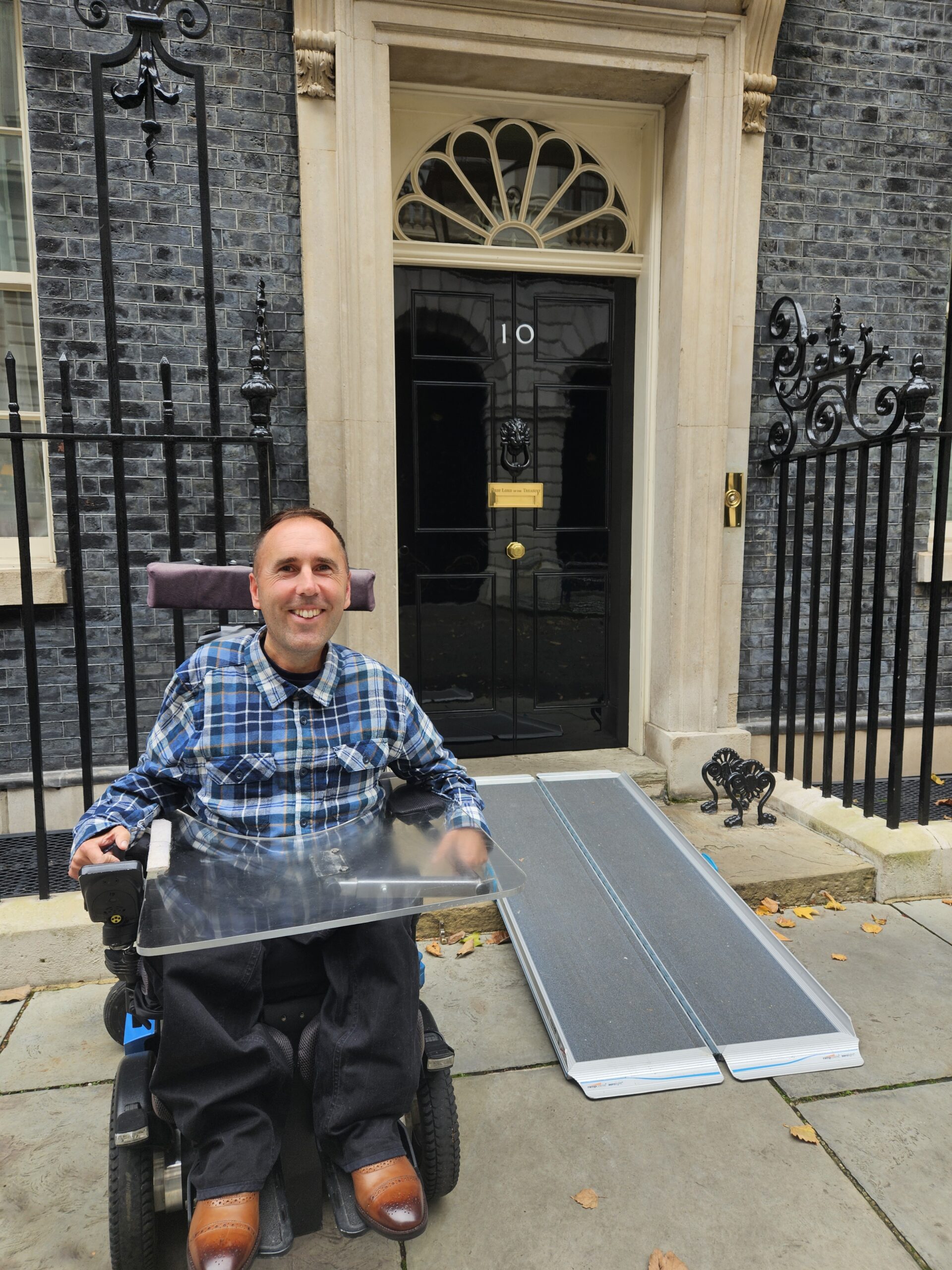 Alt text: Martyn Sibley, sitting in his wheelchair, smiles in front of the famous black door of 10 Downing Street. A ramp is laid out in front of the entrance, highlighting the building's accessibility for wheelchair users. Martyn is wearing a blue and black plaid shirt and dark trousers, with the iconic address plate and lion's head door knocker visible behind him.