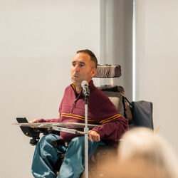 In this image, Martyn Sibley, a disability rights advocate and CEO of Purple Goat, is speaking passionately at an event. He is seated in his wheelchair, positioned at a podium with a microphone. Martyn is wearing a striped maroon and yellow sweater, engaging the audience with his insights on inclusion and accessibility. The background shows a light-filled, modern conference space, and some audience members are partially visible, attentively listening to his talk.