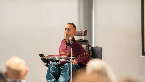 In this image, Martyn Sibley, a disability rights advocate and CEO of Purple Goat, is speaking passionately at an event. He is seated in his wheelchair, positioned at a podium with a microphone. Martyn is wearing a striped maroon and yellow sweater, engaging the audience with his insights on inclusion and accessibility. The background shows a light-filled, modern conference space, and some audience members are partially visible, attentively listening to his talk.