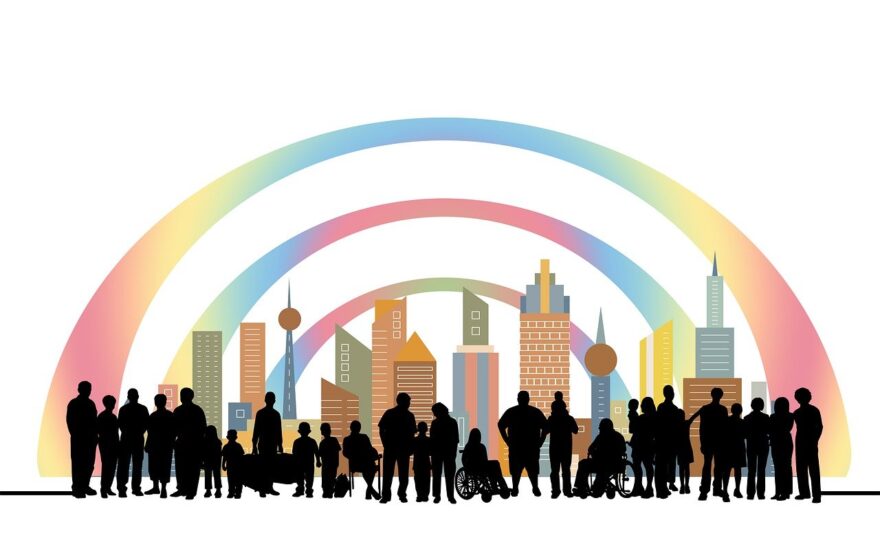 An illustration featuring a diverse group of people, including individuals with different disabilities, in front of a colorful city skyline. The group includes people of varying ages, with visible wheelchairs and other mobility aids, standing under three rainbow-colored arcs that curve above the city. The image represents inclusivity and diversity.