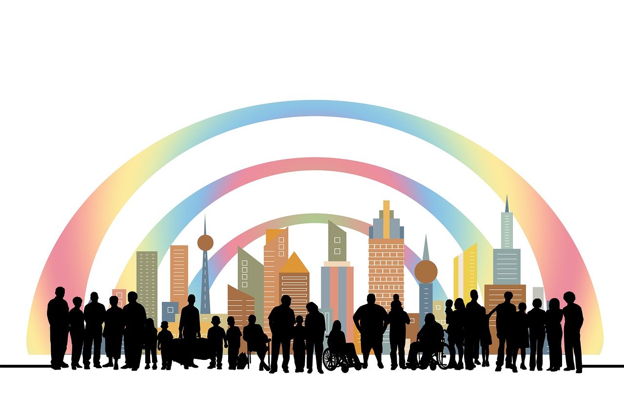 An illustration featuring a diverse group of people, including individuals with different disabilities, in front of a colorful city skyline. The group includes people of varying ages, with visible wheelchairs and other mobility aids, standing under three rainbow-colored arcs that curve above the city. The image represents inclusivity and diversity.