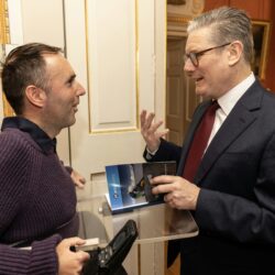 Martyn Sibley in a purple jumper, seated in his wheelchair, speaking to the Prime Minister, Sir Keir Starmer, in an elegant room with golden details on the walls. Martyn is gifting a copy of his travel book while the Prime Minister listens attentively, mid-conversation. The atmosphere is warm and professional.