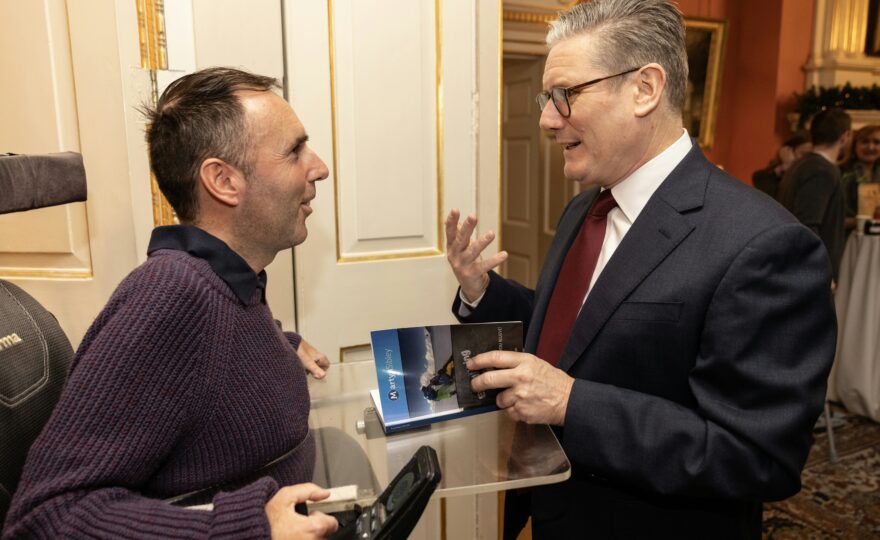 Martyn Sibley in a purple jumper, seated in his wheelchair, speaking to the Prime Minister, Sir Keir Starmer, in an elegant room with golden details on the walls. Martyn is gifting a copy of his travel book while the Prime Minister listens attentively, mid-conversation. The atmosphere is warm and professional.