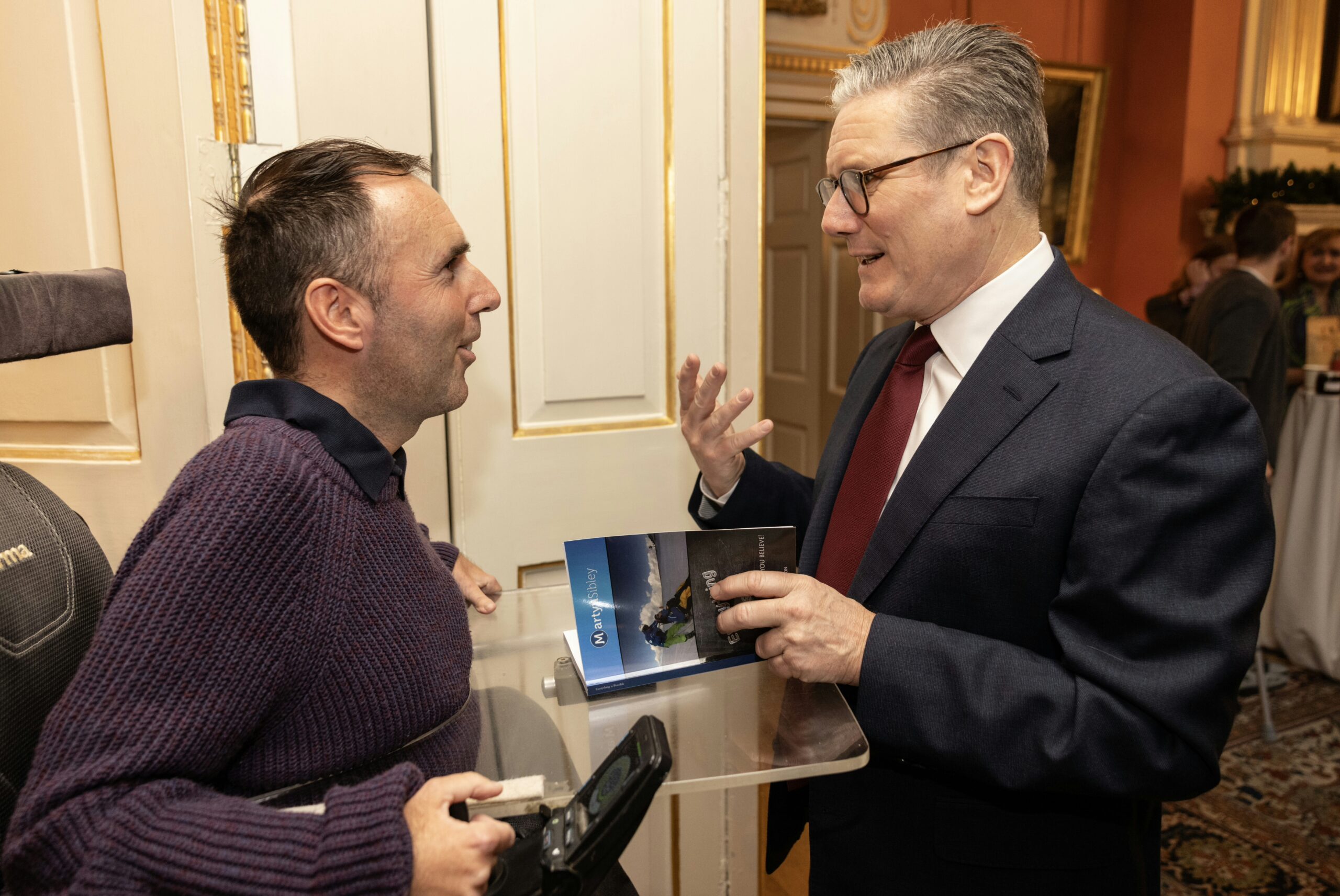 Martyn Sibley in a purple jumper, seated in his wheelchair, speaking to the Prime Minister, Sir Keir Starmer, in an elegant room with golden details on the walls. Martyn is gifting a copy of his travel book while the Prime Minister listens attentively, mid-conversation. The atmosphere is warm and professional.