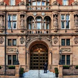 A grand, ornate facade of the Kimpton Fitzroy London hotel, showcasing intricate architectural details, statues, and a central arched entrance with a doorman standing at the top of the steps. The vibrant red blur of a passing double-decker bus emphasizes the hotel's iconic London setting. This was where I stayed last summer and used their excellent stair lift for wheelchair access.