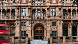 A grand, ornate facade of the Kimpton Fitzroy London hotel, showcasing intricate architectural details, statues, and a central arched entrance with a doorman standing at the top of the steps. The vibrant red blur of a passing double-decker bus emphasizes the hotel's iconic London setting. This was where I stayed last summer and used their excellent stair lift for wheelchair access.