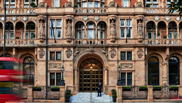 A grand, ornate facade of the Kimpton Fitzroy London hotel, showcasing intricate architectural details, statues, and a central arched entrance with a doorman standing at the top of the steps. The vibrant red blur of a passing double-decker bus emphasizes the hotel's iconic London setting. This was where I stayed last summer and used their excellent stair lift for wheelchair access.