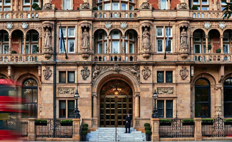 A grand, ornate facade of the Kimpton Fitzroy London hotel, showcasing intricate architectural details, statues, and a central arched entrance with a doorman standing at the top of the steps. The vibrant red blur of a passing double-decker bus emphasizes the hotel's iconic London setting. This was where I stayed last summer and used their excellent stair lift for wheelchair access.