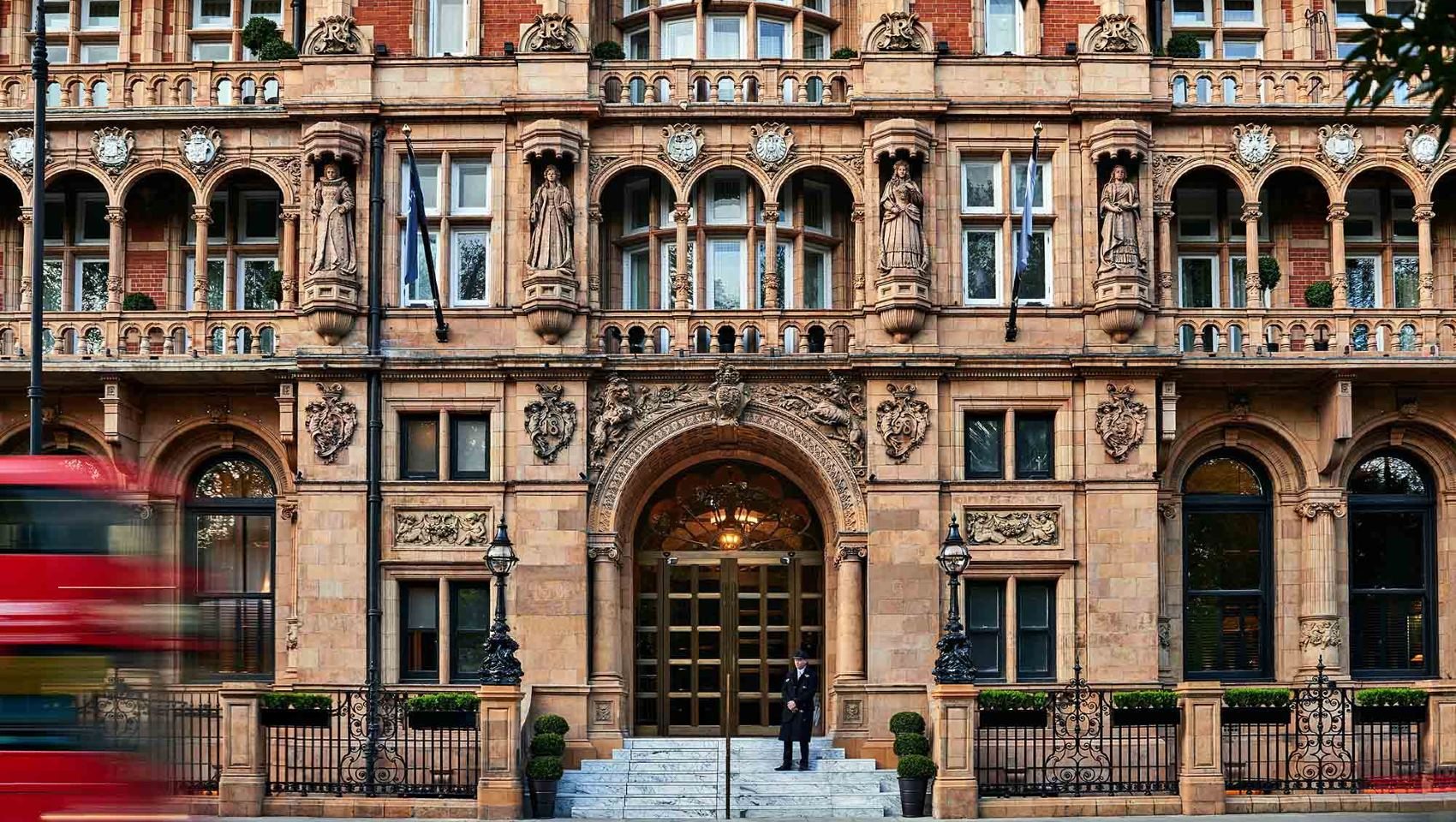 A grand, ornate facade of the Kimpton Fitzroy London hotel, showcasing intricate architectural details, statues, and a central arched entrance with a doorman standing at the top of the steps. The vibrant red blur of a passing double-decker bus emphasizes the hotel's iconic London setting. This was where I stayed last summer and used their excellent stair lift for wheelchair access.