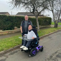 Martyn is seated in his brand-new purple Permobil F5 wheelchair, smiling with Ray standing beside him. The wheelchair features a transparent tray. They are outdoors on a quiet residential street with neatly trimmed hedges, leafless trees, and brick houses in the background. Ray played a key role in the assessment and delivery of the chair.