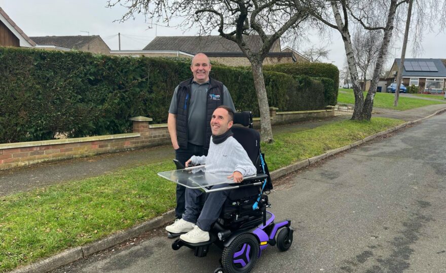 Martyn is seated in his brand-new purple Permobil F5 wheelchair, smiling with Ray standing beside him. The wheelchair features a transparent tray. They are outdoors on a quiet residential street with neatly trimmed hedges, leafless trees, and brick houses in the background. Ray played a key role in the assessment and delivery of the chair.