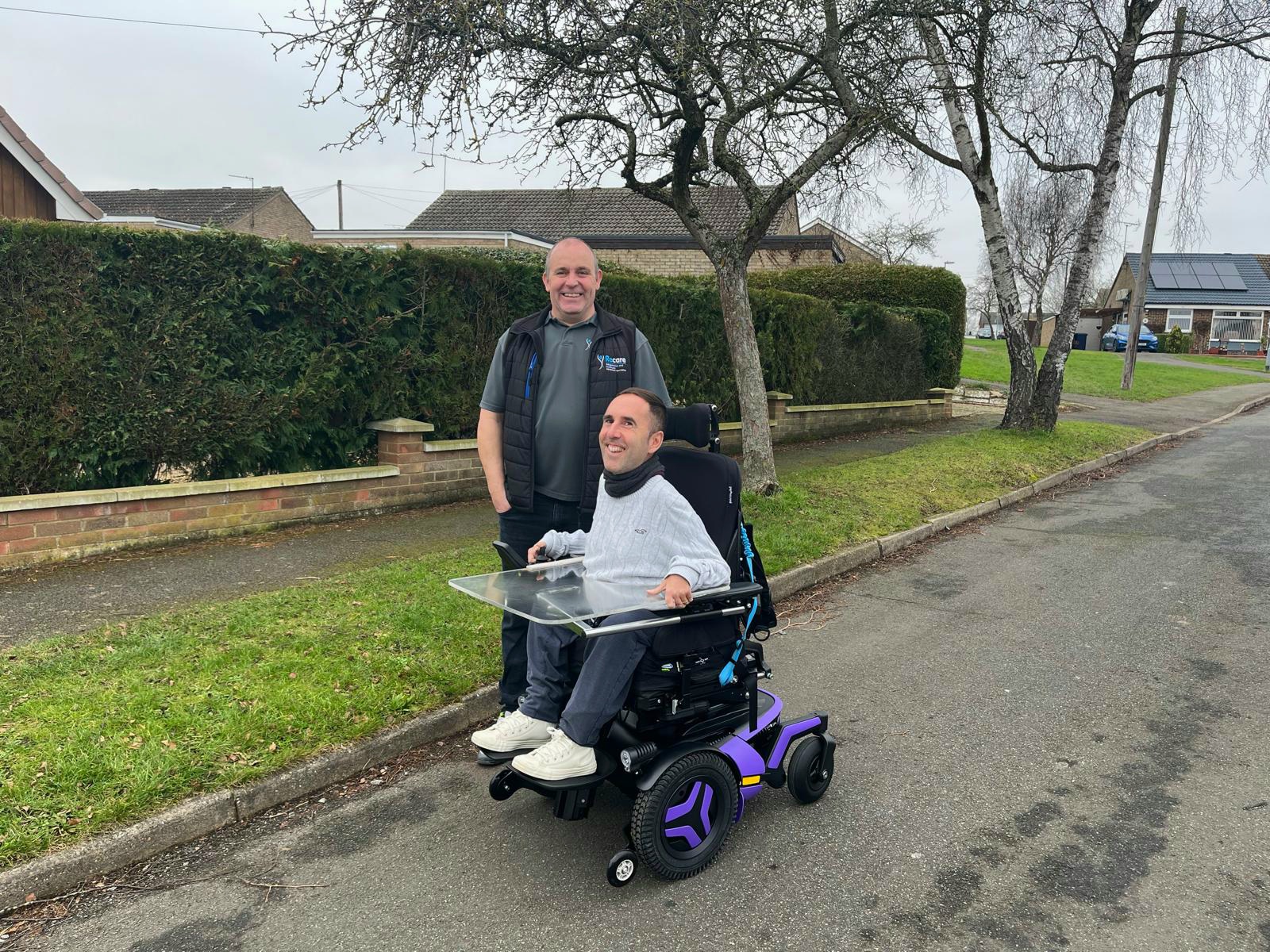 Martyn is seated in his brand-new purple Permobil F5 wheelchair, smiling with Ray standing beside him. The wheelchair features a transparent tray. They are outdoors on a quiet residential street with neatly trimmed hedges, leafless trees, and brick houses in the background. Ray played a key role in the assessment and delivery of the chair.