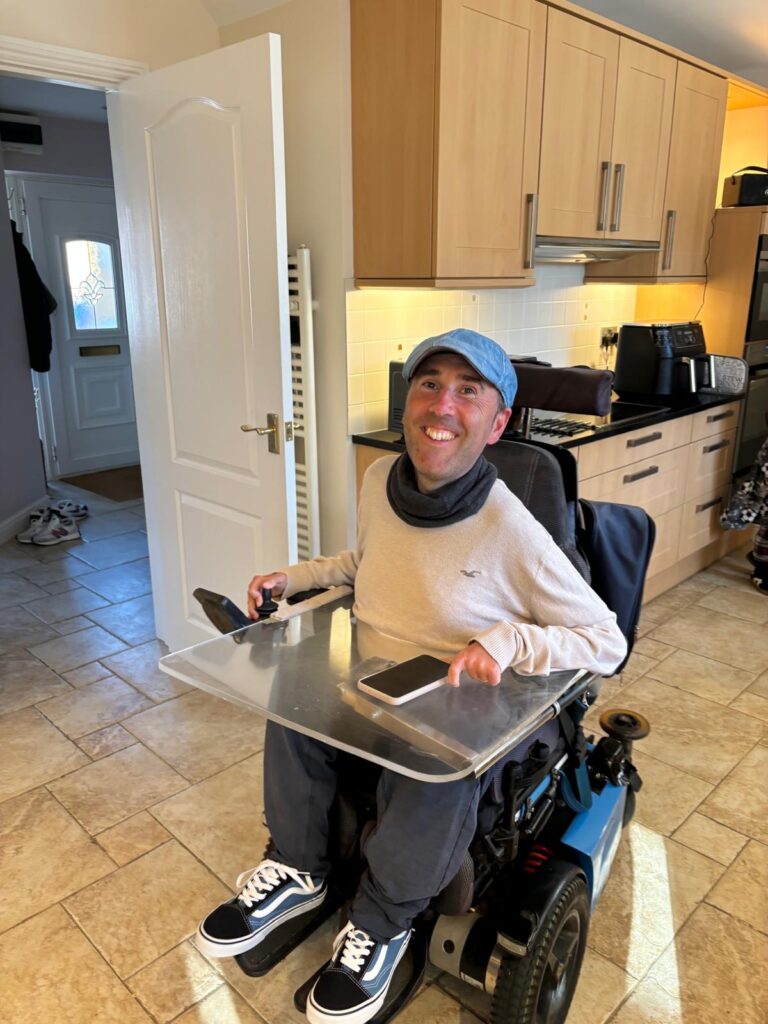 Martyn Sibley smiling in a bright kitchen, wearing a light jumper, navy trousers, blue cap, and Vans trainers, sitting in his powered wheelchair with a clear tray attached, holding his phone.