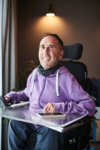 Martyn Sibley smiling warmly while sitting in his powered wheelchair, wearing a purple hoodie. He is indoors, with soft lighting above his head and a plant in the background. A smartphone rests on his transparent tray.