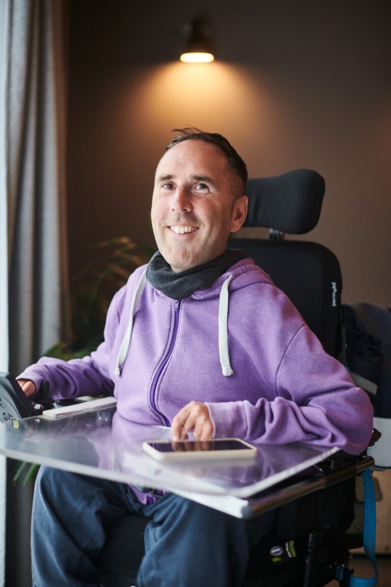 Martyn Sibley smiling warmly while sitting in his powered wheelchair, wearing a purple hoodie. He is indoors, with soft lighting above his head and a plant in the background. A smartphone rests on his transparent tray.