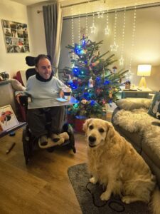Martyn sits in his powered wheelchair beside a decorated Christmas tree, smiling with a drink on his tray. His golden retriever Sunny sits on the floor nearby, looking at the camera. Warm fairy lights, cosy living room, festive and relaxed atmosphere.