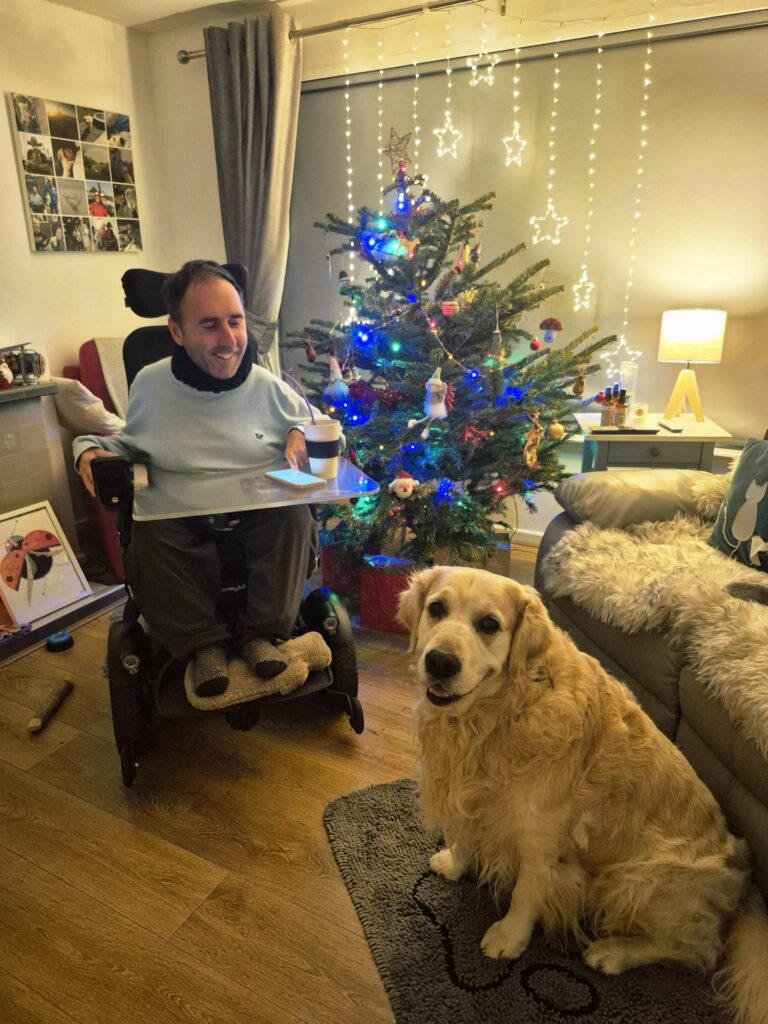 Martyn sits in his powered wheelchair beside a decorated Christmas tree, smiling with a drink on his tray. His golden retriever Sunny sits on the floor nearby, looking at the camera. Warm fairy lights, cosy living room, festive and relaxed atmosphere.