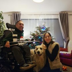 Martyn sits in his powered wheelchair beside his wife Kasia and their golden retriever Sunny in a cosy living room. They are gathered by a decorated Christmas tree with lights, creating a warm, relaxed end-of-year family moment.