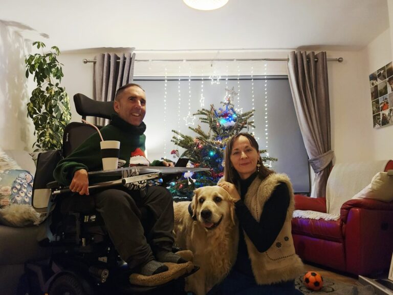 Martyn sits in his powered wheelchair beside his wife Kasia and their golden retriever Sunny in a cosy living room. They are gathered by a decorated Christmas tree with lights, creating a warm, relaxed end-of-year family moment.