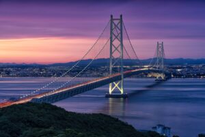 A long suspension bridge stretches across calm water at sunset, glowing with lights. The sky fades from pink to deep purple, and a city skyline sits in the background with mountains behind it.