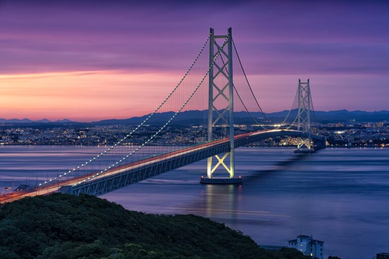 A long suspension bridge stretches across calm water at sunset, glowing with lights. The sky fades from pink to deep purple, and a city skyline sits in the background with mountains behind it.