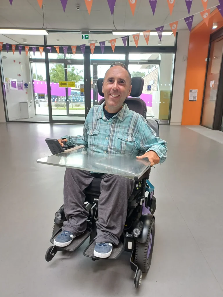 Martyn Sibley smiling in his powered wheelchair in the entrance area of the Sense building in Birmingham, wearing a blue and green checked shirt. Purple and orange Sense bunting hangs overhead, with glass entrance doors behind him. Photo taken during a visit for a Sense trustee board meeting.